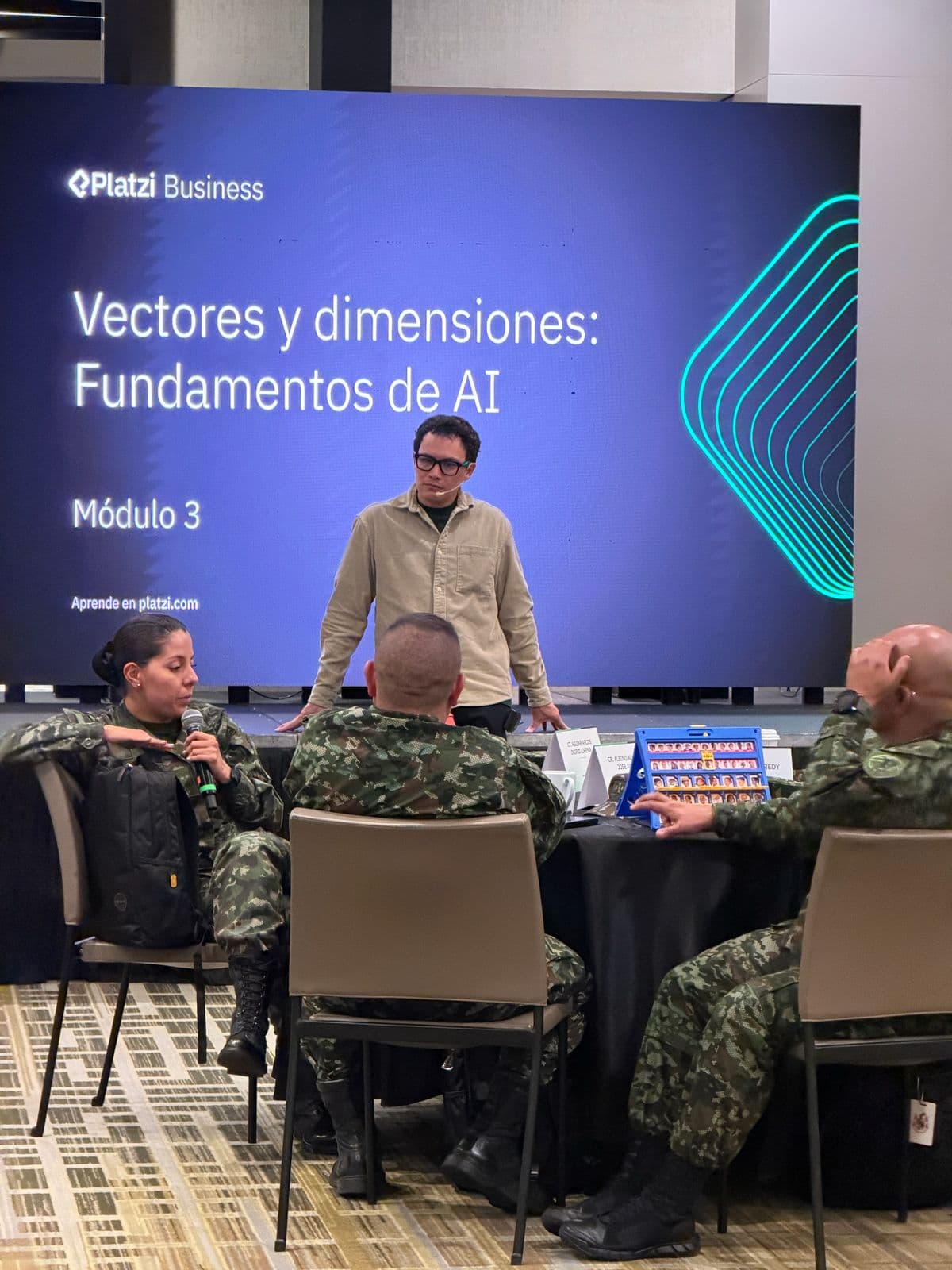 Fabián Oloarte facilitating a corporate AI session in front of a screen titled Vectores y dimensiones: Fundamentos de AI while participants discuss at tables.