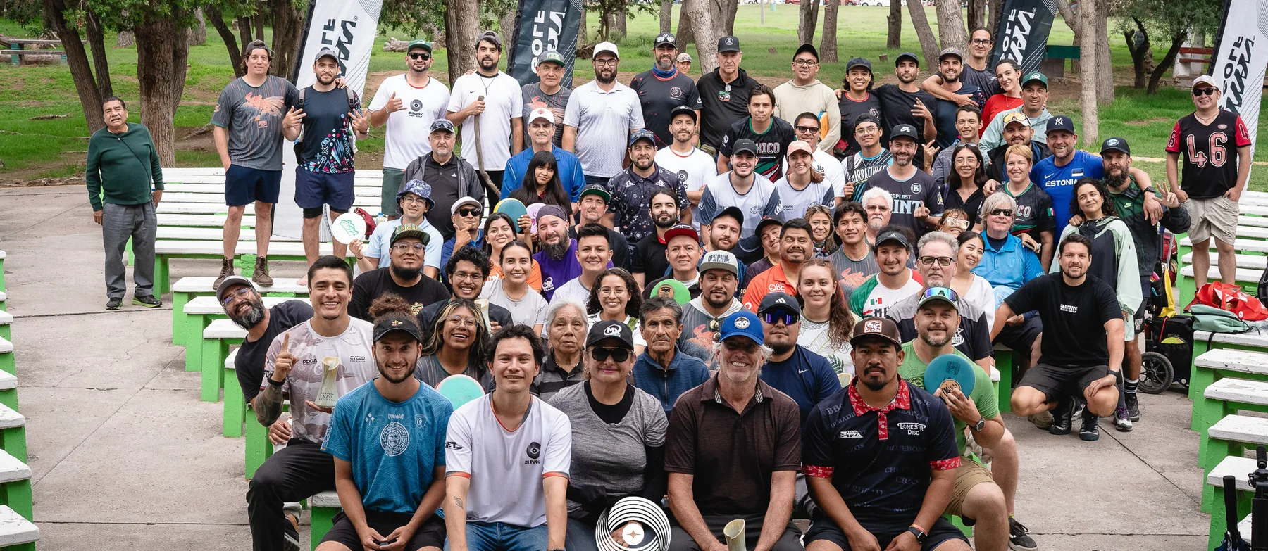 A large disc golf community gathered for a group photograph in a wooded park.